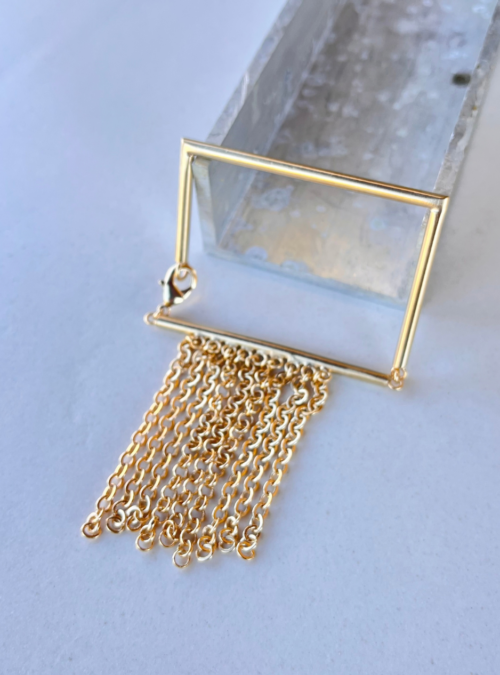 Bracciale a forma rettangolare in ottone costruito in filo con snodo e chiusura a moschettone e frangia di catene. Gioielli particolari dal colore dell'oro chiaro satinato